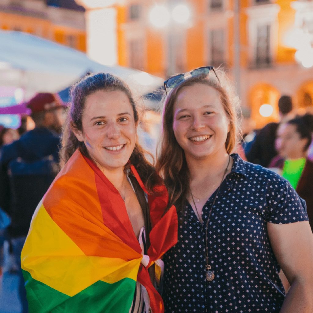Portrait of two smiling girls in Lisbon. One of them is wrapped around a LGBTQIA+ flag.
