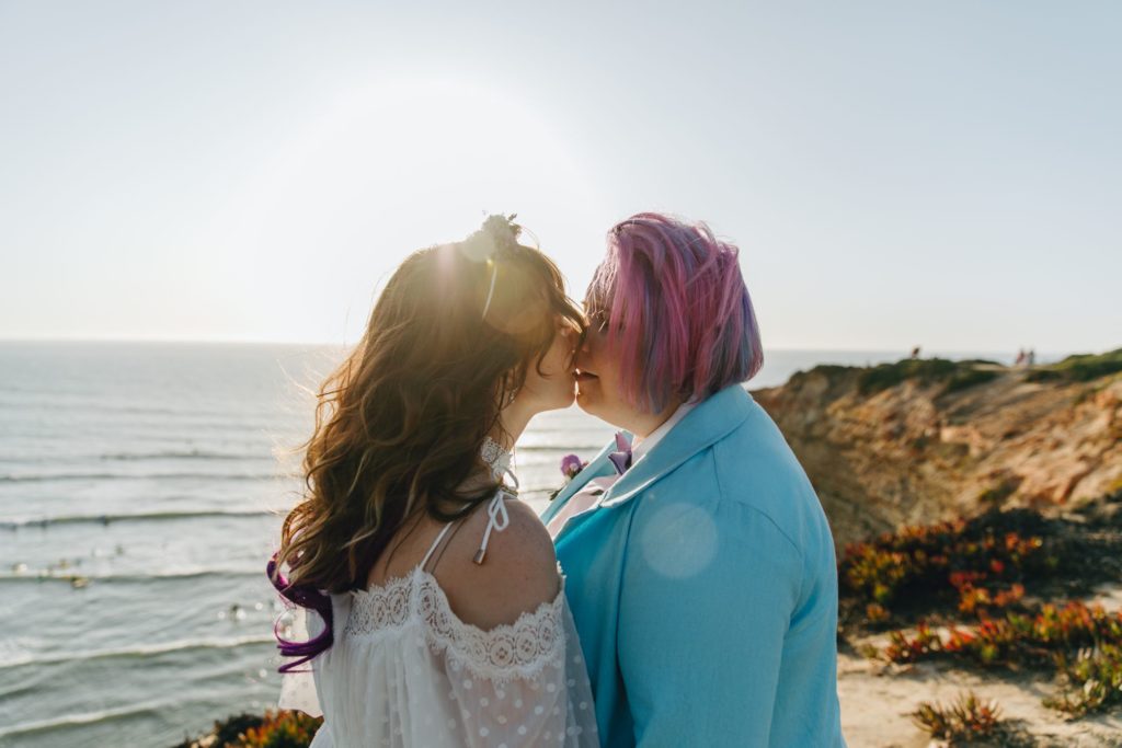 Couple of two women kissing each other with a view to a beach in Portugal. One of the woman is wearing a blue blazer, while the other is wearing a white lace dress.