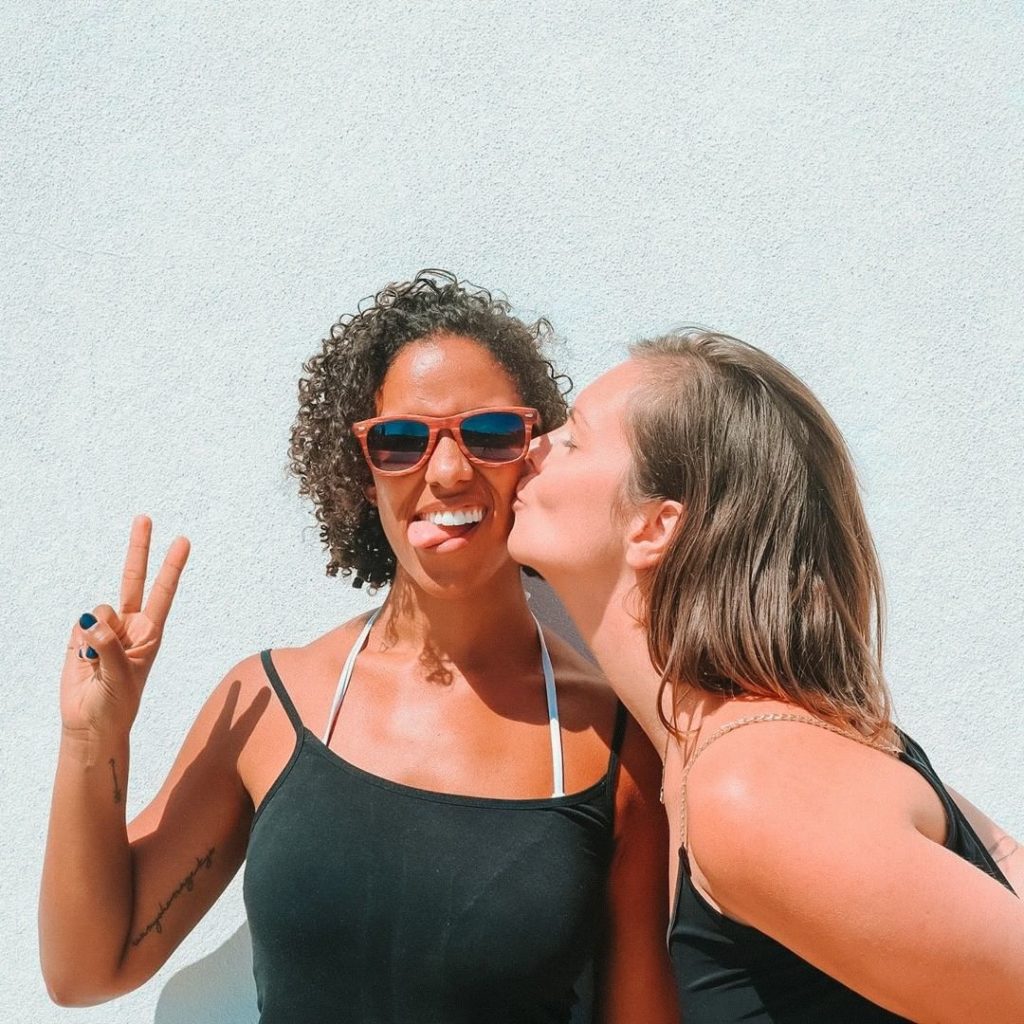 Two girls in black tank tops. One of them is kissing the other on the cheek, while the other is making the “peace sign”.