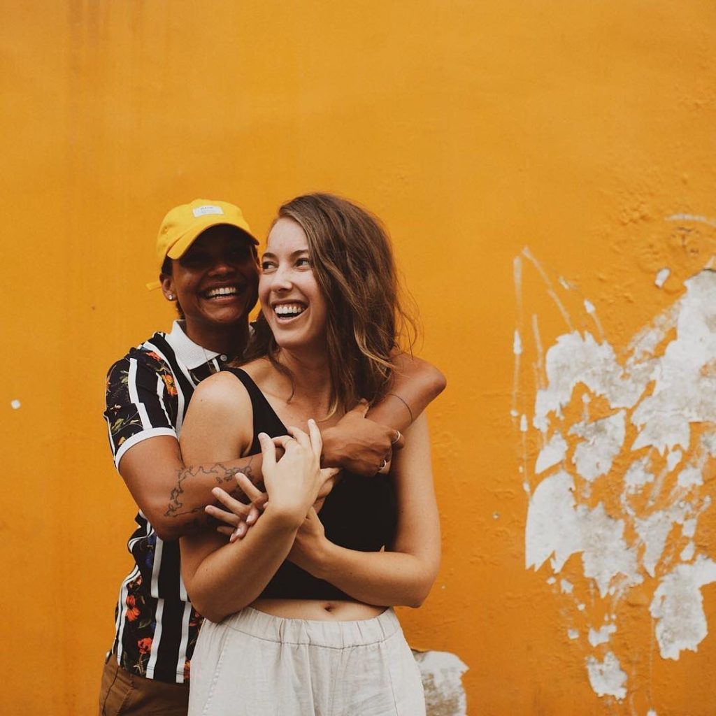 Two girls, one of them hugging the other from behind. In the background there’s a yellow wall.