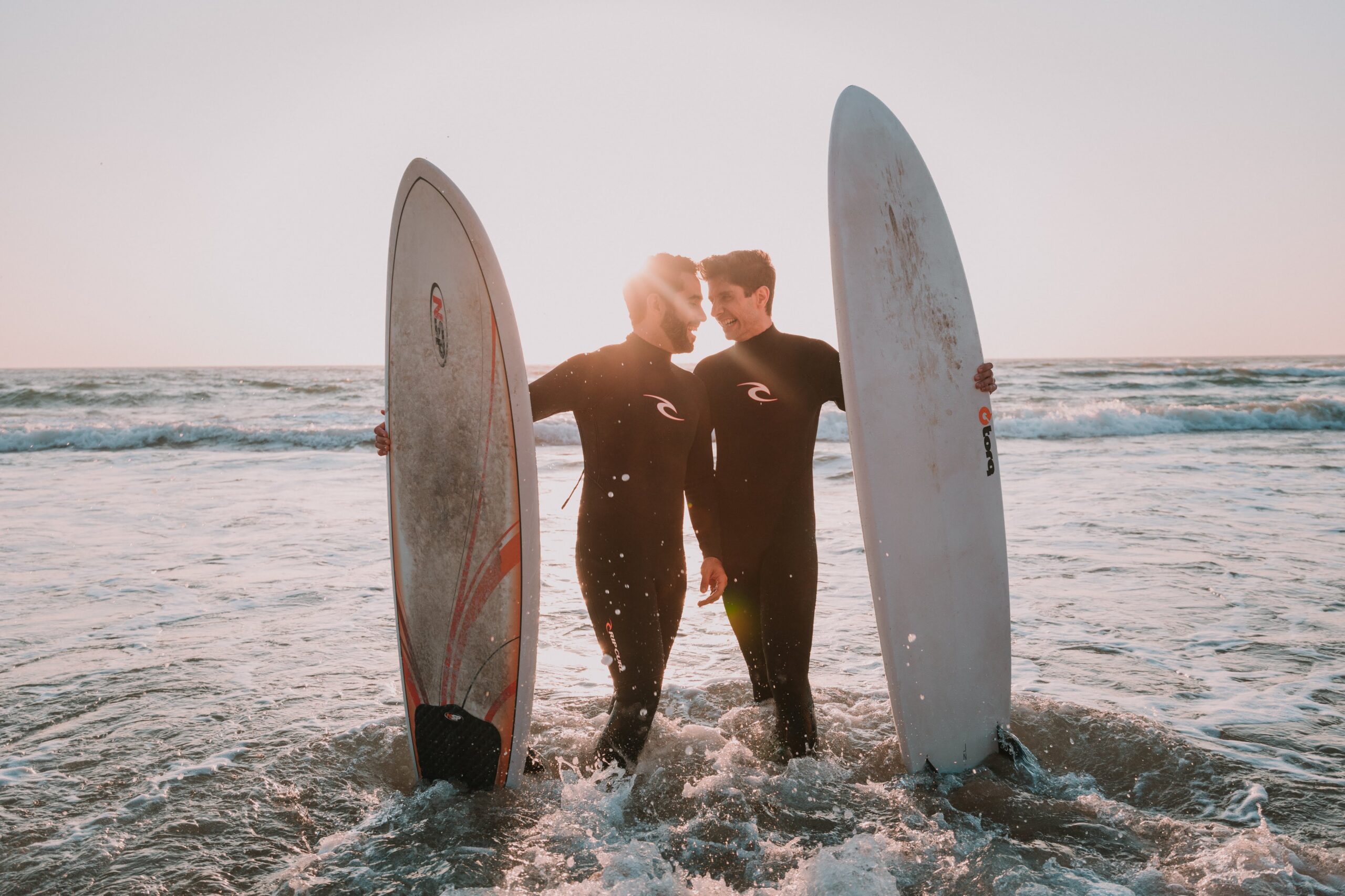 Two men smiling at each other and holding surf boards at the beach