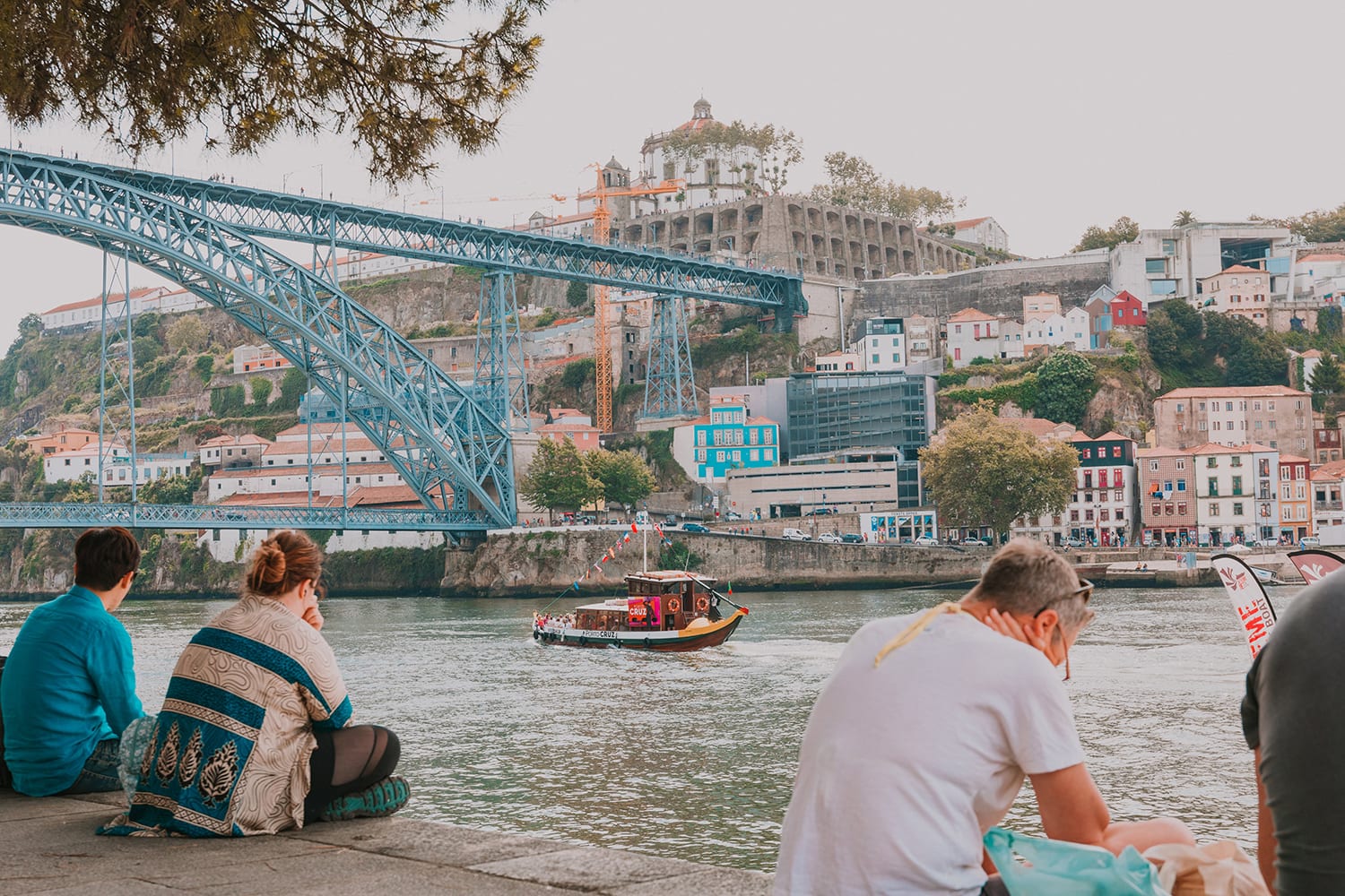 People sitting on the floor with a view to D. Luís I Bridge. In the river there's a small boat passing by.